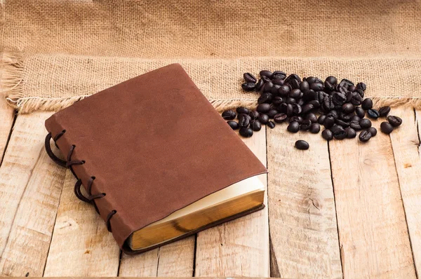 Verstreute Schwarze Kaffeebohnen Und Geschlossenes Notizbuch Auf Holzgrund — Stockfoto