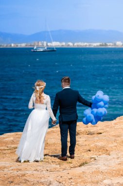 Beyaz gelinlik içinde gelin ve koyu mavi takım elbiseli damat, kayalıklarda el ele tutuşuyorlar, güzel mavi deniz arka planına karşı. Damat mavi hava balonları tutuyor. Düğün Mallorca, 