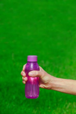 Eko plastiğin mavi su şişesini tutan el çimlerin üzerinde duruyor. Su için ekolojik plastik kavanoz. Çevre koruma kavramı, doğa bakımı, plastiksiz çevre. Boşluğu kopyala.