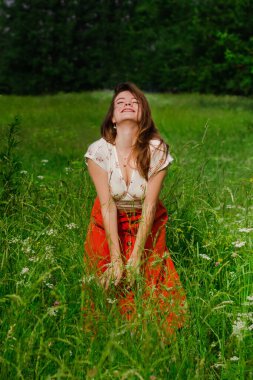Beyaz bluzlu, kırmızı etekli, yeşil çayır çimenlerde dikilen, saçlarına dokunan ve kırsal hayatın ve doğanın tadını çıkaran genç esmer kızın yumuşak odağı.