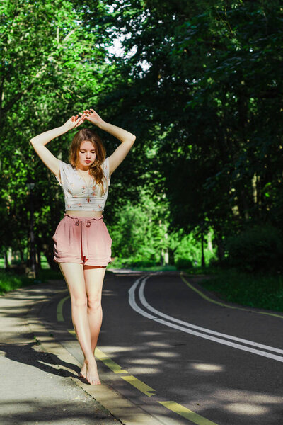 Young woman alone in pink shorts and white top with hands up walking and dancing barefoot in the middle of drive way in the park looking down. Solo outdoor activity. End quarantine