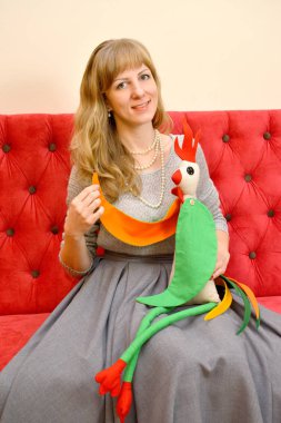 Kırmızı koltuk elinde oyuncak horoz ile genç güzel kadın oturur