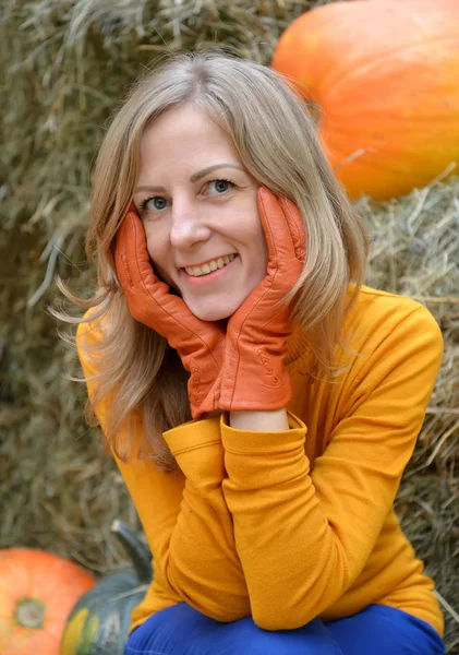 Une jeune femme étreint son visage avec ses mains en gants orange. Portrait sur fond de citrouille et de foin — Photo