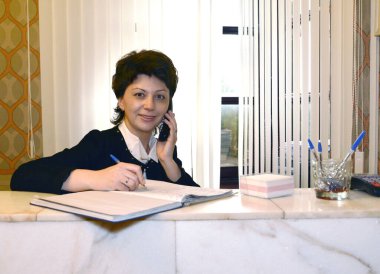 Otel yöneticisi telefonda konuşuyor ve kasanın arkasındaki kütüğü dolduruyor.