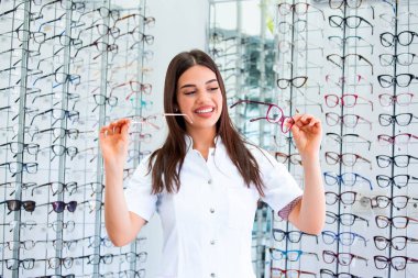 Attractive young female doctor in ophthalmology clinic. Doctor ophthalmologist is standing near shelves with different eyeglasses. clipart