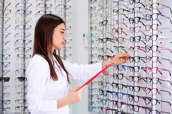 Ελκυστική γυναίκα οφθαλμίατρο γράφοντας δεδομένα σε ένα πρόχειρο, δουλεύοντας σε ένα οπτικό κατάστημα. Έννοια της υγειονομικής περίθαλψης και της ιατρικής — Φωτογραφία Αρχείου