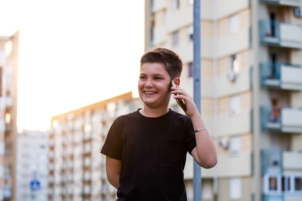 Tiener jongen glimlachend tijdens het praten met zijn vrienden via sociale netwerken met behulp van mobiele telefoon, zittend tegen stedelijke landschap achtergrond — Stockfoto