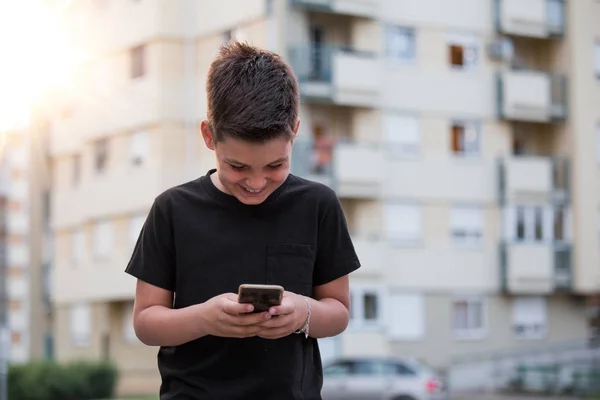 Ragazzo adolescente sorridente mentre invia messaggi ai suoi amici tramite i social network utilizzando il telefono cellulare, seduto sullo sfondo del paesaggio urbano — Foto Stock