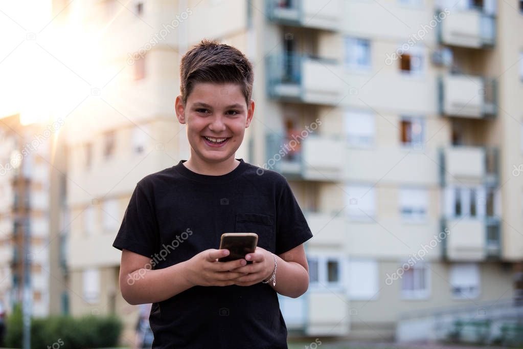 Teenage boy in street wear looking and smiling at the camera with cheerful expression while texting his friends via social networks using mobile phone, sitting against urban landscape background