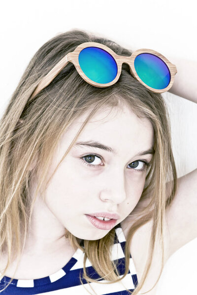Blond girl eleven years old standing near white wall with green sunglasses