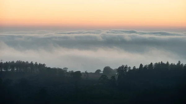 Epic landscape image of cloud inversion at sunset over Dartmoor National Park in Engand with cloud rolling through forest on horizon