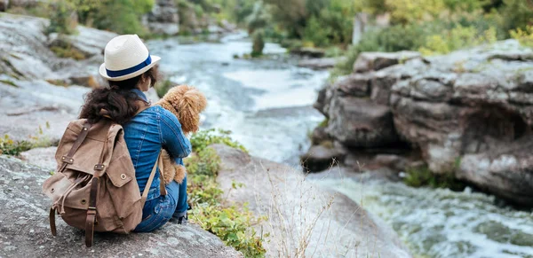 Woman traveler with backpack holding dog and looking at natural canyon with view of the mountain river.