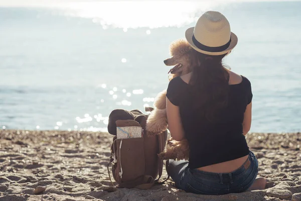 Woman traveler with backpack holding dog and looking at sea. Concept of travel.