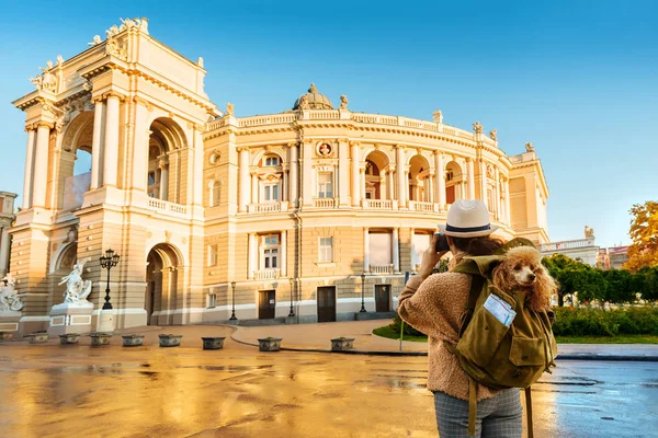 Woman traveler with dog in the backpack making pictures with a camera while traveling. Odessa Opera Theatre.