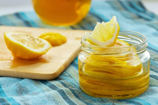 Slices of lemon, sprinkled with sugar, are in a glass jar. Nearby, on a wooden cutting board, is a sliced lemon. A cup of tea is seen in the background
