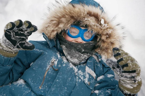 一个穿着暖和蓝色眼镜的孩子躺在雪地上看着镜头 — 图库照片