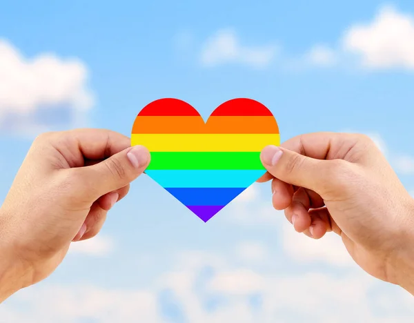 Two gay men holding a heart painted like a LGBT flag against sunny blue sky background. Lgbt flag rainbow heart shape on blue sky background.