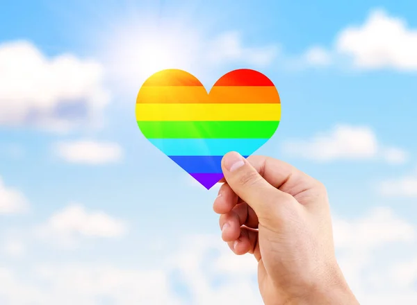 Lgbt flag rainbow heart shape on blue sky background. Man hand holding a heart painted like a LGBT flag.