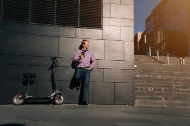 Modern iş merkezinin yanında duran akıllı telefon ve elektrikli scooterlı adam..