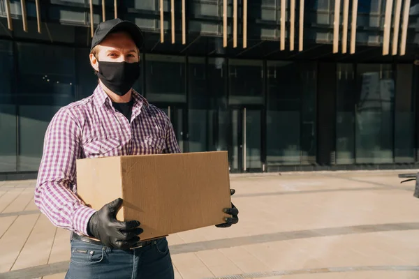 Young European man in a medical mask and black disposable gloves a cardboard paper box. The concept of contactless delivery of food from a restaurant. online stores