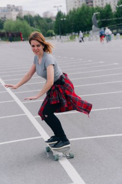 Şehirde sörf tahtasıyla gezen güzel bir genç kadın. Ekose gömlekli şık bir kız otoparkta kaykay sürüyor..