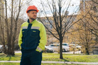 Temiz erkek müteahhit başka tarafa bakıyor. Özel elbiseli ve kask takan bir adam, arka planda ağaçların karşısında duruyor.