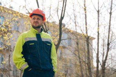 Temiz erkek müteahhit başka tarafa bakıyor. Özel elbiseli ve kask takan bir adam, arka planda ağaçların karşısında duruyor.