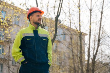 Temiz erkek müteahhit başka tarafa bakıyor. Özel elbiseli ve kask takan bir adam, arka planda ağaçların karşısında duruyor.