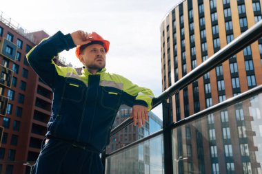 Temiz erkek müteahhit başka tarafa bakıyor. Özel elbiseli ve kasklı bir adam, bir iş merkezinin arka planına karşı planın ortasında bir ev inşa ederken görülüyor.