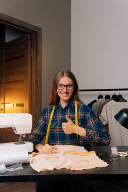 Gülümseyen moda tasarımcısı iş yerinde kameraya bakıyor, terzi, terzi ya da atölye sahibi masada oturmuş moda tasarımının taslaklarıyla, küçük iş konseptiyle