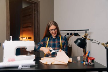 Gülümseyen bir terzi kameraya baktı ve elbisenin bir taslağını kesti. Terzi kız dikiş atölyesinde çalışıyor..