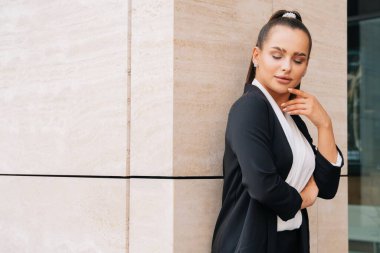 Zarif, şık bir bayan siyah bir takım elbiseyle bir iş merkezinin duvarına yaslanıyor. Kız zevkle gözlerini kapattı..