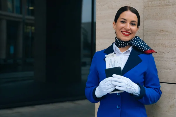 A smiling flight attendant holds a passport and a boarding pass. Portrait of a young flight attendant in uniform look into the camera. High quality photo