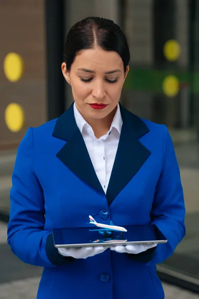 A sad flight attendant looks at a mini plane in a white gloves blue work suit. Bright flight attendant misses.