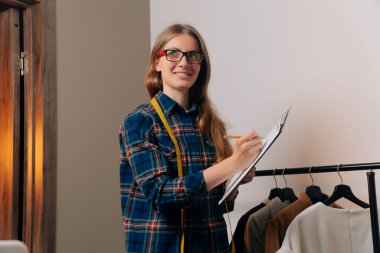 Bir atölyede dikiş ekipmanları ile kendinden emin bir terzi elinde tabletle müşterilerin ölçüsünü alıyor..