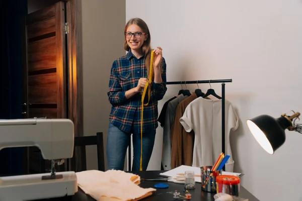 A brooding seamstress stands next to things. Tailor removes clothes on dress hangers.