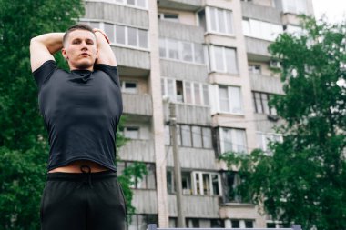 Sırt kaslarını esneten genç bir vücut geliştiricinin portresi. Sokaktaki apartmanının arka planına karşı..