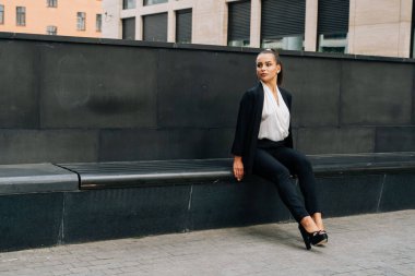 Siyah takım elbiseli bir iş kadını öğle yemeği saatinde iş yerinin dışındaki bankta oturuyor. Müşteri bekleme konsepti.