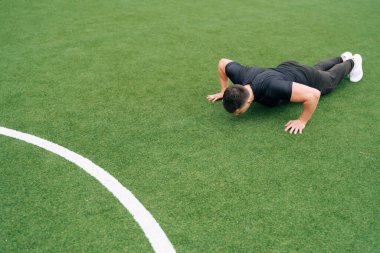 Atletik adam stadyumun oyun alanında yeşil çimlerde şınav çekiyor. Aktif spor kavramı sadece koronavirüs tarafından belirlenir..
