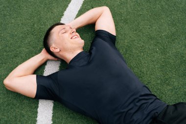 Gülümseyen bir sporcunun portresi futbol sahasındaki yeşil çimlerin üzerinde yatıyor. Açık hava eğitiminin yorgunluk kavramı.