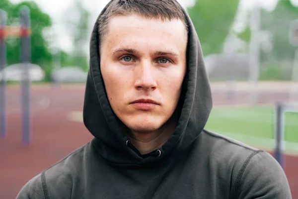 Portrait of a sportsman in a black hooded suit against the backdrop of a sports field, the treadmill empty