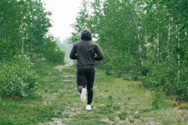 Siyah eşofmanlı bir adam parkta ormana doğru koşuyor. Sabah koşusu konsepti, dikiz aynası atletizm koçu.