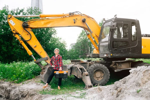 Woman in construction clothes with a white helmet in a plaid shirt on the background of an excavator. Engineer concept at a construction site next to pit excavation equipment