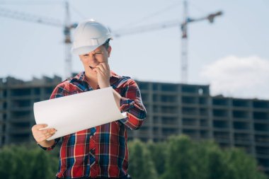 Beyaz koruyucu kasklı bir adam portresi. Elinde bir bina inşaatı kağıdıyla. Doğanın mavi gökyüzüne sahip bir vincin arka planındaki akıllı mühendis kavramı..