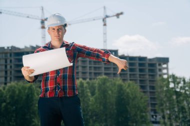 Beyaz kasklı üzgün bir adamın portresi. Elinde binanın arka planında kağıt var. Başparmağını aşağıda tut..