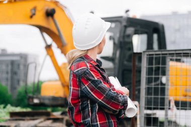 Kazıcının arka planında beyaz kasklı, ekose gömlekli bir kadın. Çukur kazma ekipmanlarının yanındaki inşaat sahasında mühendis kavramı