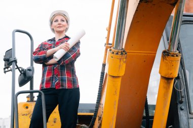 Kazıcının arka planında beyaz kasklı, ekose gömlekli bir kadın. Çukur kazma ekipmanlarının yanındaki inşaat sahasında mühendis kavramı