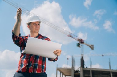 Beyaz koruyucu kasklı bir adam portresi. Elinde bir bina inşaatı kağıdıyla. Doğanın mavi gökyüzüne sahip bir vincin arka planındaki akıllı mühendis kavramı..