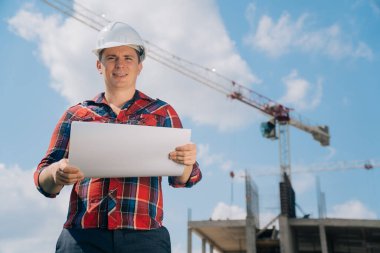 Beyaz koruyucu kasklı bir adam portresi. Elinde bir bina inşaatı kağıdıyla. Doğanın mavi gökyüzüne sahip bir vincin arka planındaki akıllı mühendis kavramı..