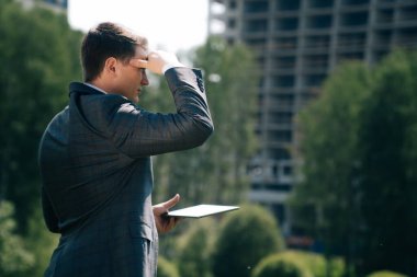 Şık bir genç adam şehrin arka planına karşı dijital bir tablet tutuyor. İnşaat mühendisi kavramının yanında çok katlı bir bina inşaatı ile ilgili belgeler bulunmakta.